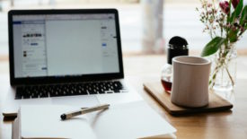 Laptop and notebook open on wood desk next to a coffee cup